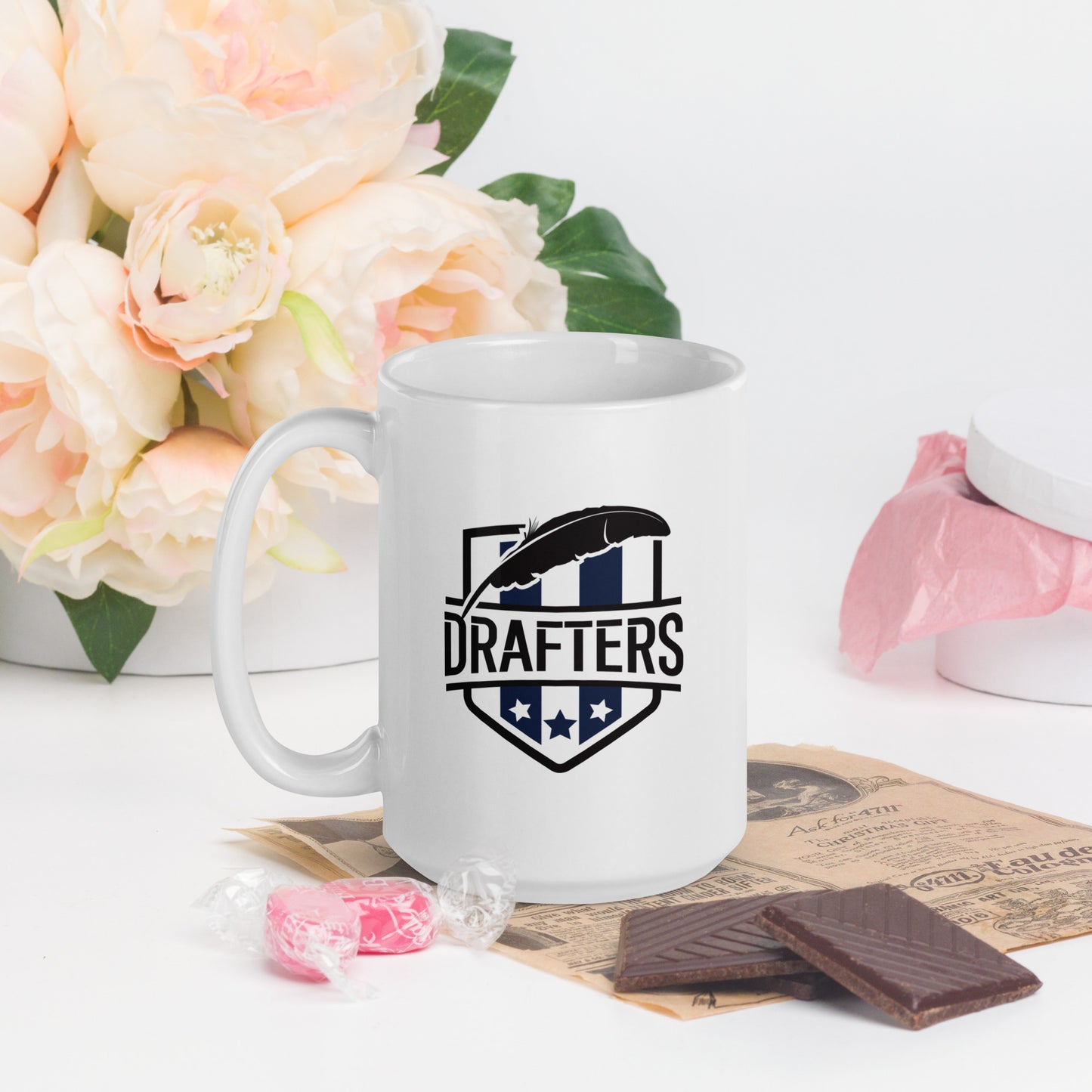 A white coffee mug with a school crest on it, prominently featuring the word Drafters as the team name, with a large quill over top of the letters