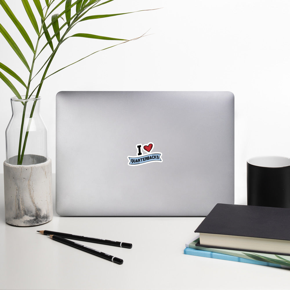 A sticker that reads "I Heart Quarterbacks" with a large red heart, Quarterbacks is printed on a blue banner. The sticker is 3 x 3 inches