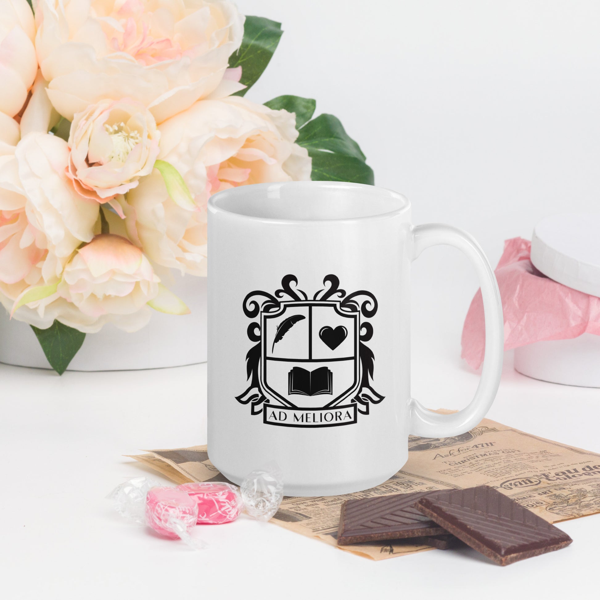 A white mug with a coat of arms logo. The coat of arms features an open book, a quill, a heart, and the words Ad Meliora below