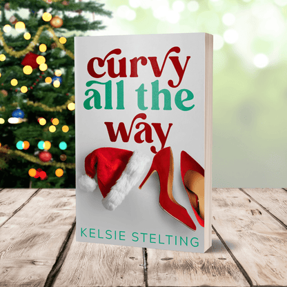 An paperback cover sits on a table, with christmas lights behind it. The book is titled Curvy All the Way by Kelsie Stelting and features a santa hat and red heels on the cover
