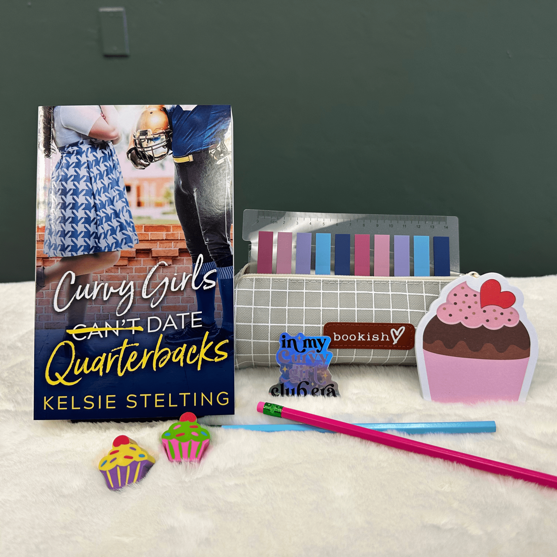 A book cover showing a plus size woman next to a football player reads Curvy Girls Can't Date Quarterbacks, the word Can't is crossed out. Authored by Kelsie Stelting. The book sits on a table next to pens, cupcake stickers of varying sizes and other pieces of memorabilia 