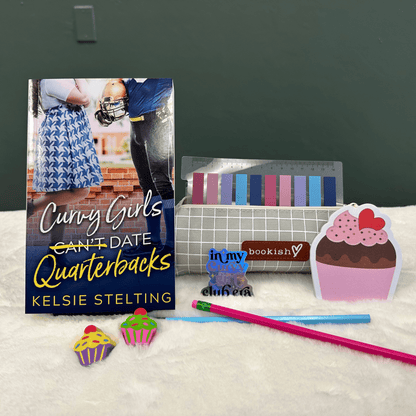 A book cover showing a plus size woman next to a football player reads Curvy Girls Can't Date Quarterbacks, the word Can't is crossed out. Authored by Kelsie Stelting. The book sits on a table next to pens, cupcake stickers of varying sizes and other pieces of memorabilia 