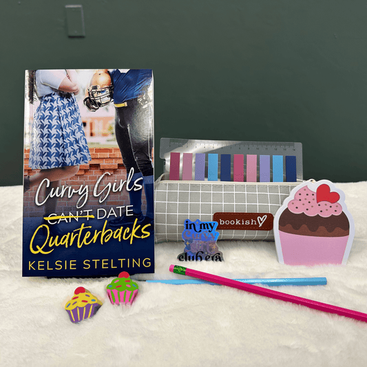 A book cover showing a plus size woman next to a football player reads Curvy Girls Can't Date Quarterbacks, the word Can't is crossed out. Authored by Kelsie Stelting. The book sits on a table next to pens, cupcake stickers of varying sizes and other pieces of memorabilia 