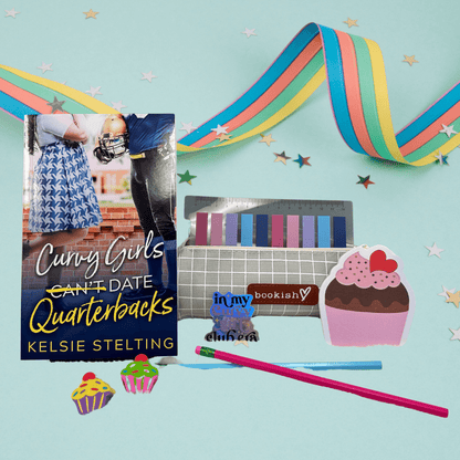 A book cover showing a plus size woman next to a football player reads Curvy Girls Can't Date Quarterbacks, the word Can't is crossed out. Authored by Kelsie Stelting. The book sits on a table next to pens, cupcake stickers of varying sizes and other pieces of memorabilia 