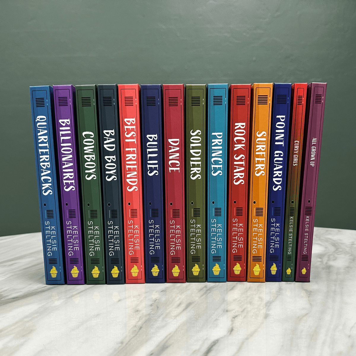 A stack of books on a table, with titles visible on the back binding. In order from left to right they read: Quarterbacks, Billionaires, Cowboys, Bad Boys, Best Friends, Bullies, Dance, Soldiers, Princes, Rockstars, Surfers, Point Guards, Curvy Girls, and All Grown Up 
