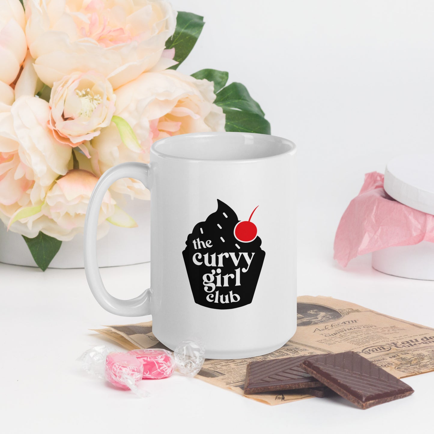 A white coffee mug featuring a black cupcake logo, with white words inside that read "the Curvy Girl Club"