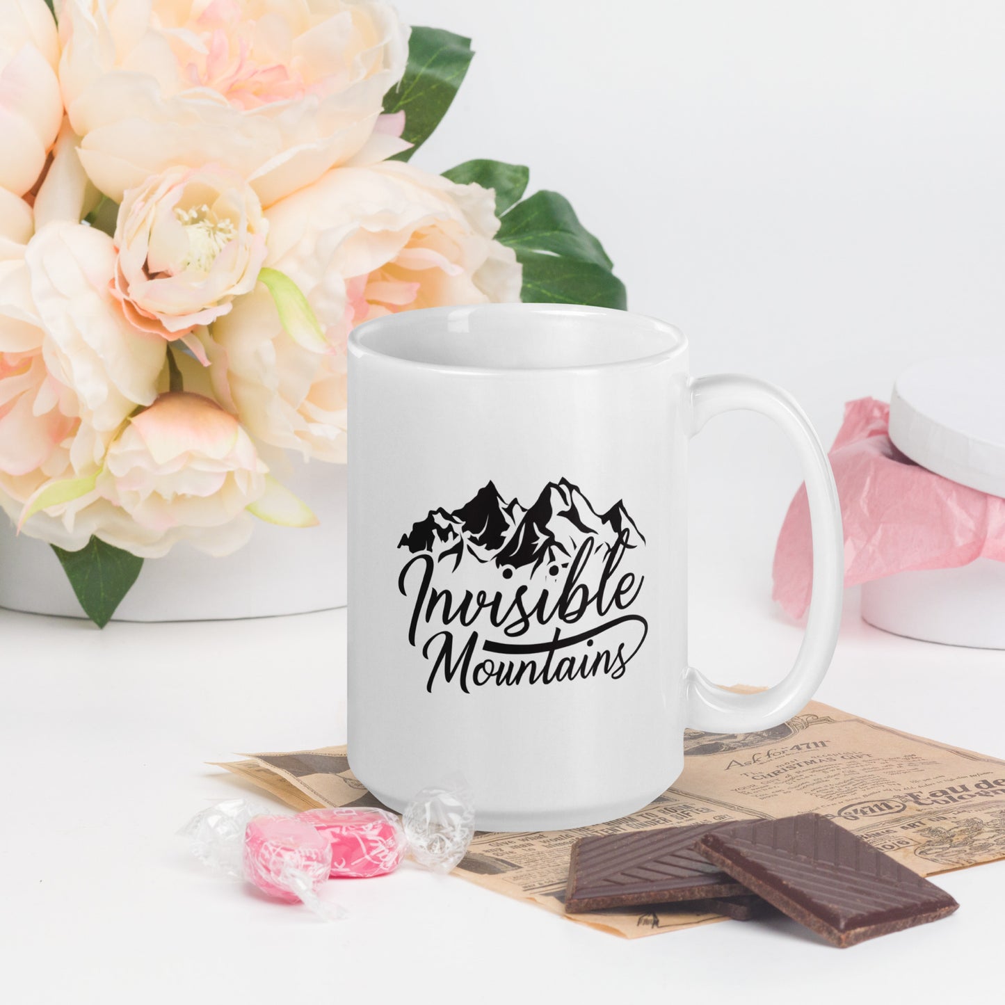 A white coffee mug with the Invisible Mountains Logo. The Invisible Mountains Logo shows the words Invisible Mountain written in cursive under a stylized mountain range 