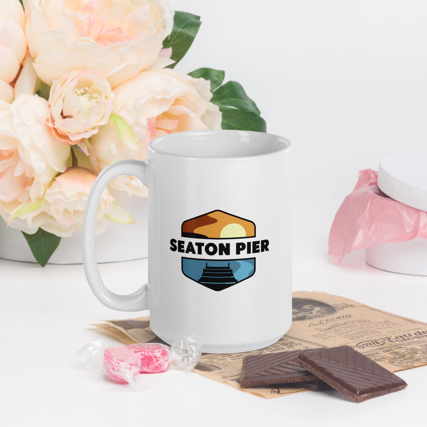 A white coffee mug with the Seaton Pier logo. The Seaton pier logo features the words Seaton Pier separating the sun and the ocean, with a dark pier on the water