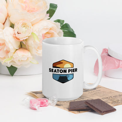 A white coffee mug with the Seaton Pier logo. The Seaton pier logo features the words Seaton Pier separating the sun and the ocean, with a dark pier on the water