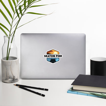 A sticker that shows a colorful setting sun over an ocean pier. With the words Seaton Pier in the middle, printed on a sticker that is 5.55.5 inches