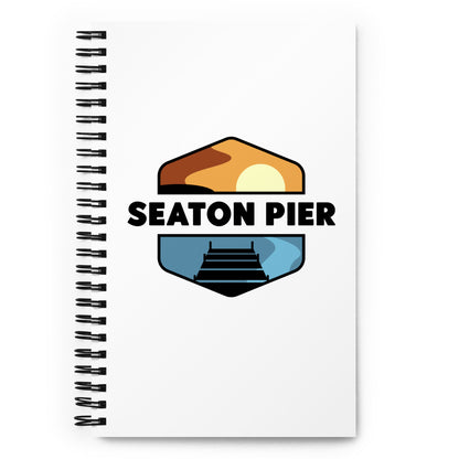 a notebook with the Seaton Pier logo on the cover. The Seaton pier logo features the words Seaton Pier separating the sun and the ocean, with a dark pier on the water
