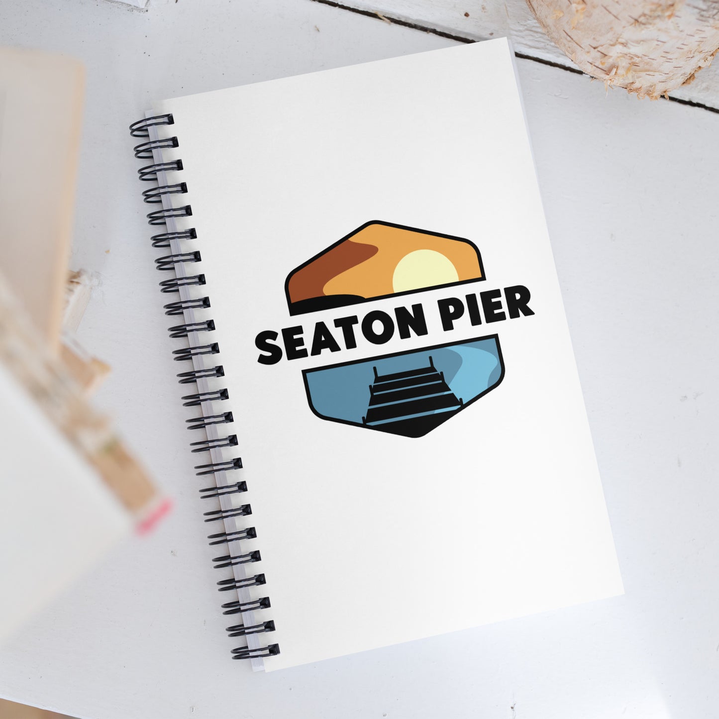 a notebook with the Seaton Pier logo on the cover. The Seaton pier logo features the words Seaton Pier separating the sun and the ocean, with a dark pier on the water