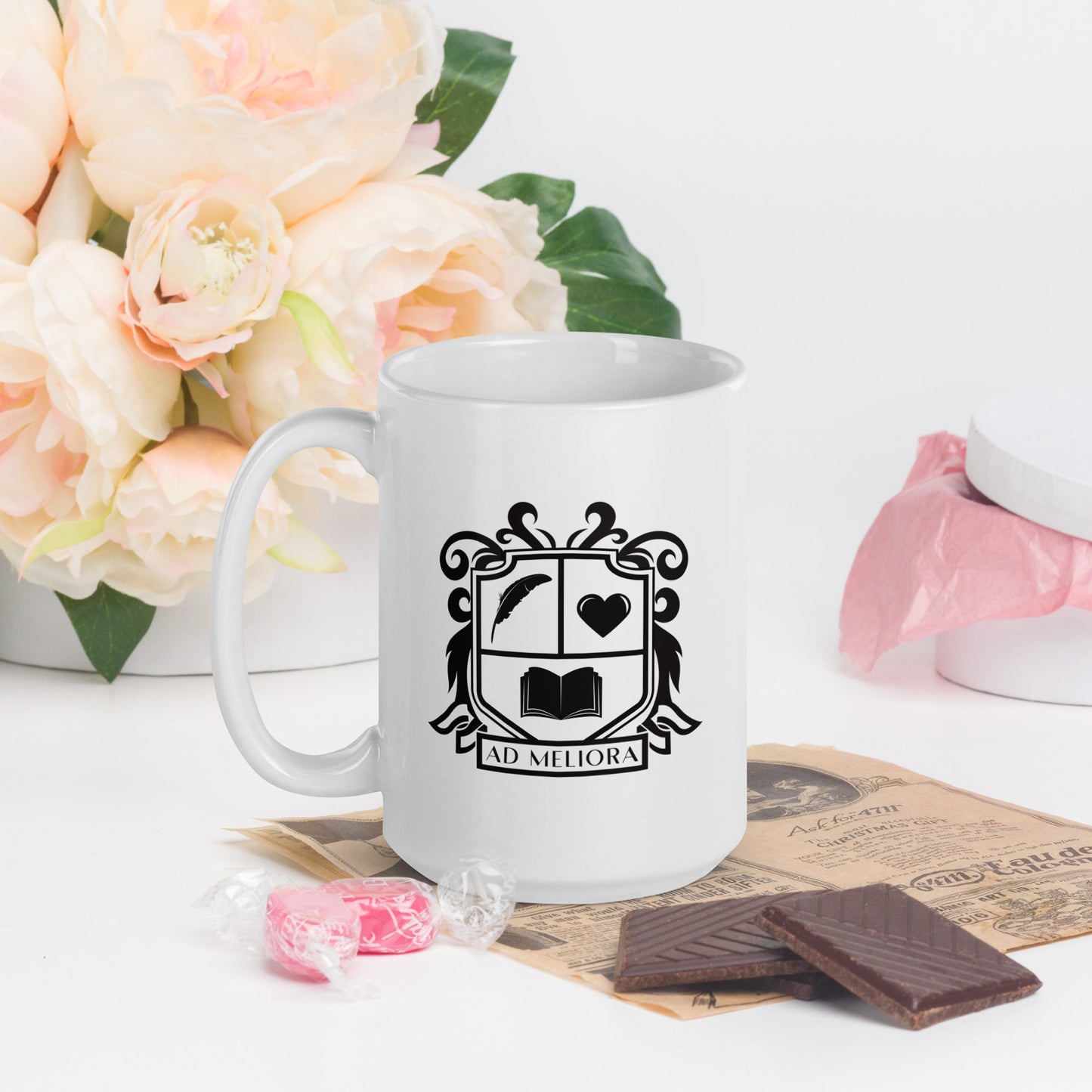 A white mug with a coat of arms logo. The coat of arms features an open book, a quill, a heart, and the words Ad Meliora below