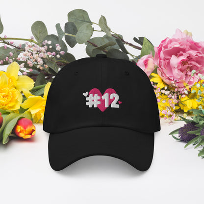 a black cap with the I Heart #12 Logo. The Number 12 Logo is a large pink heart with #12 in black written over the heart.