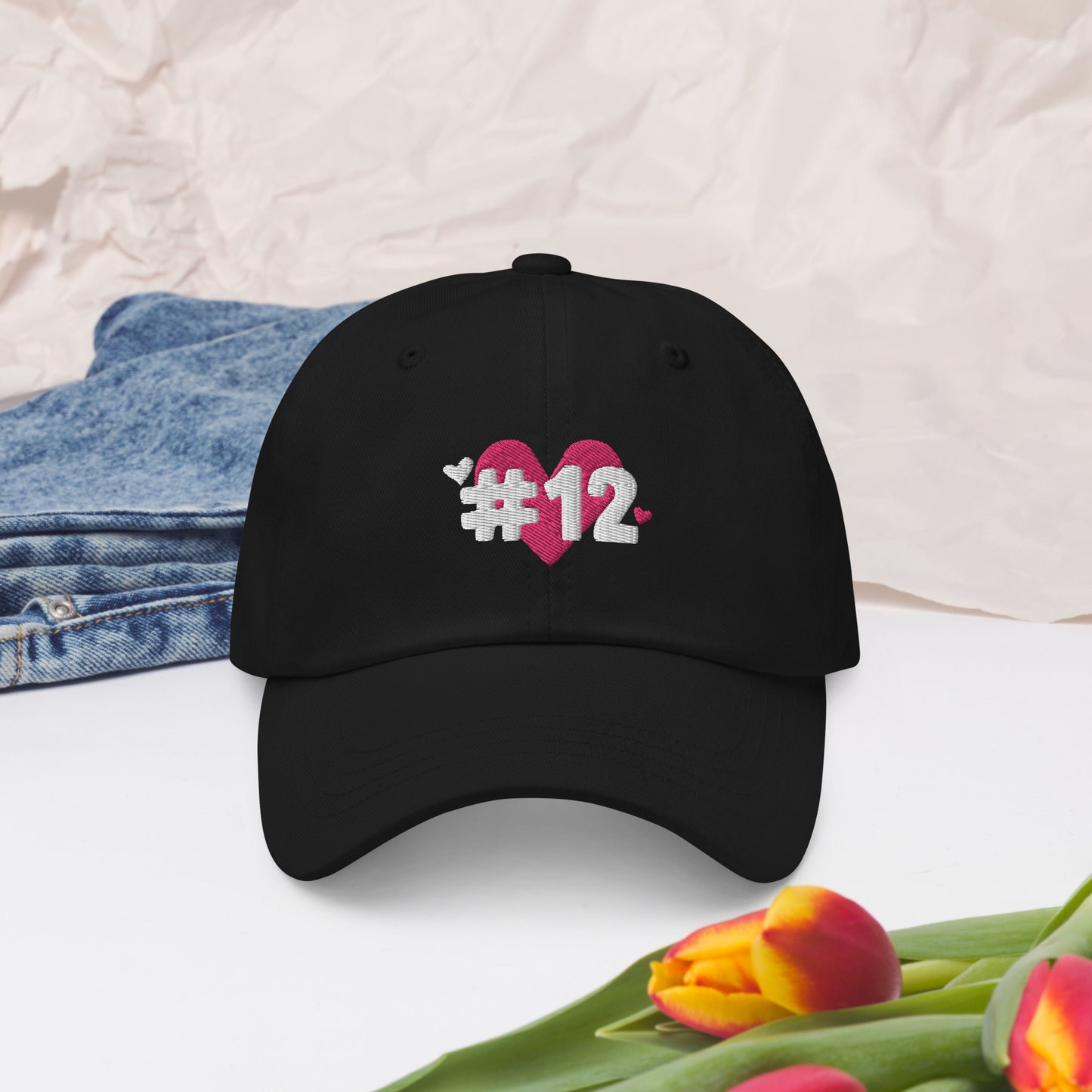a black cap with the I Heart #12 Logo. The Number 12 Logo is a large pink heart with #12 in black written over the heart.