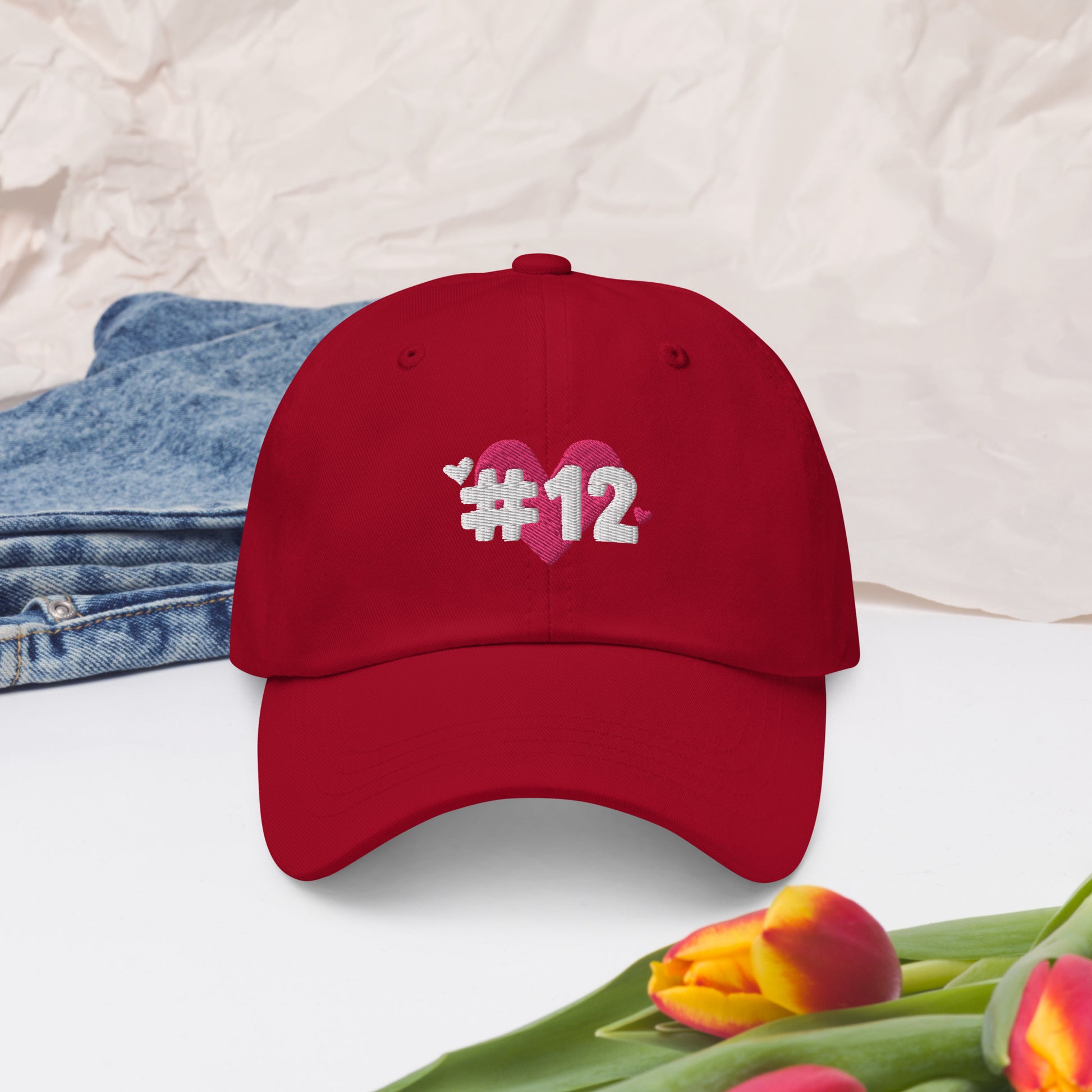 a cranberry cap with the I Heart #12 Logo. The Number 12 Logo is a large pink heart with #12 in black written over the heart.