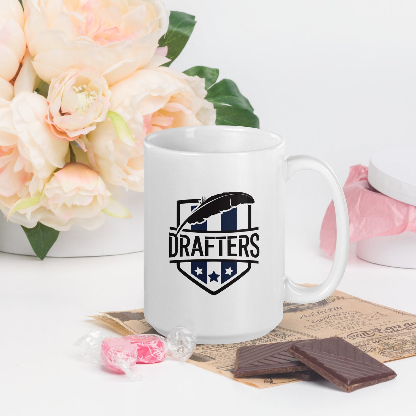 A white coffee mug with a school crest on it,  prominently featuring the word Drafters as the team name, with a large quill over top of the letters