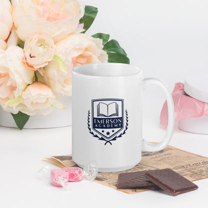 A white mug with the Emerson Academy logo, stylized like a school crest, prominently featuring an open book