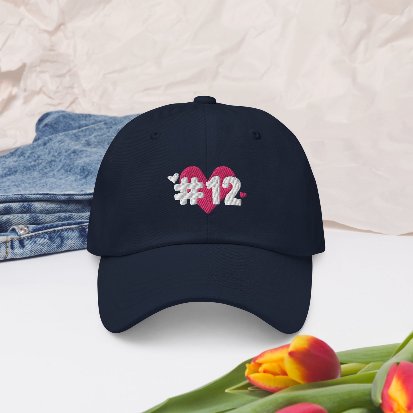 a navy cap with the I Heart #12 Logo. The Number 12 Logo is a large pink heart with #12 in black written over the heart.