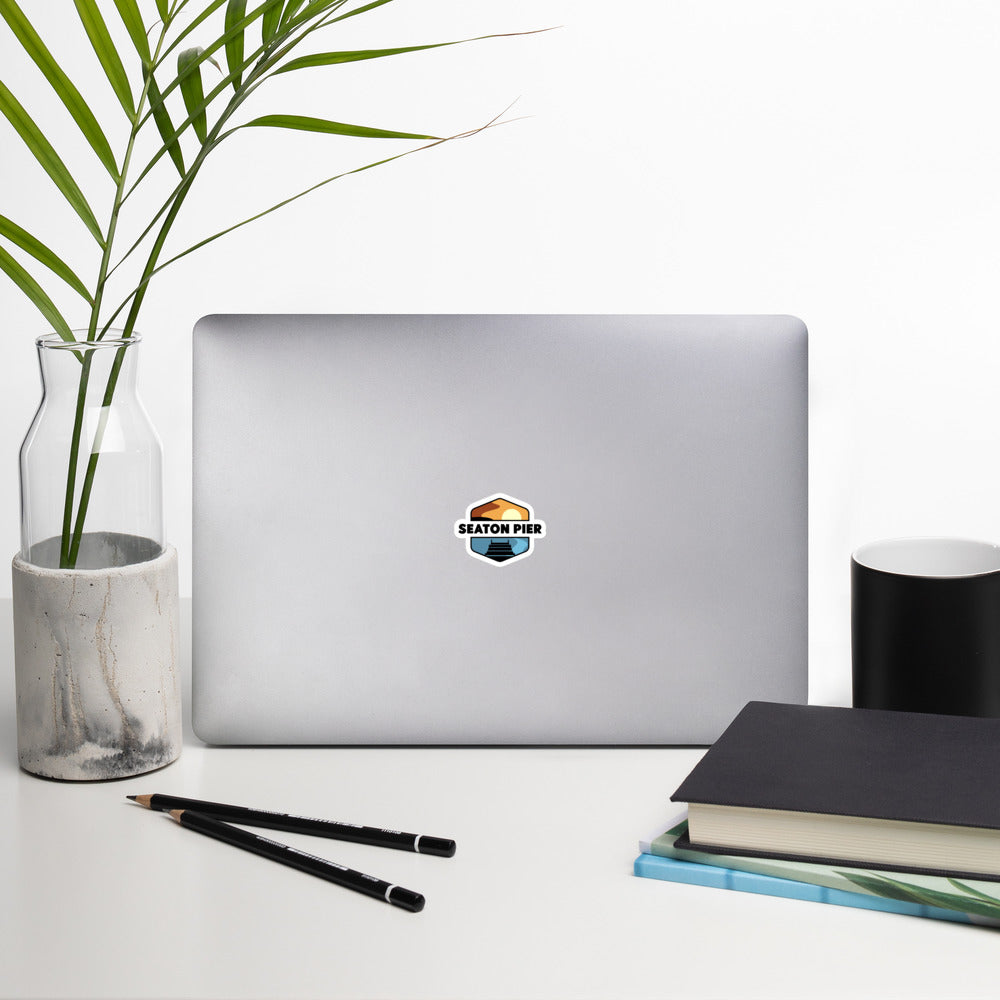 A sticker that shows a colorful setting sun over an ocean pier. With the words Seaton Pier in the middle, printed on a sticker that is 3x3 inches