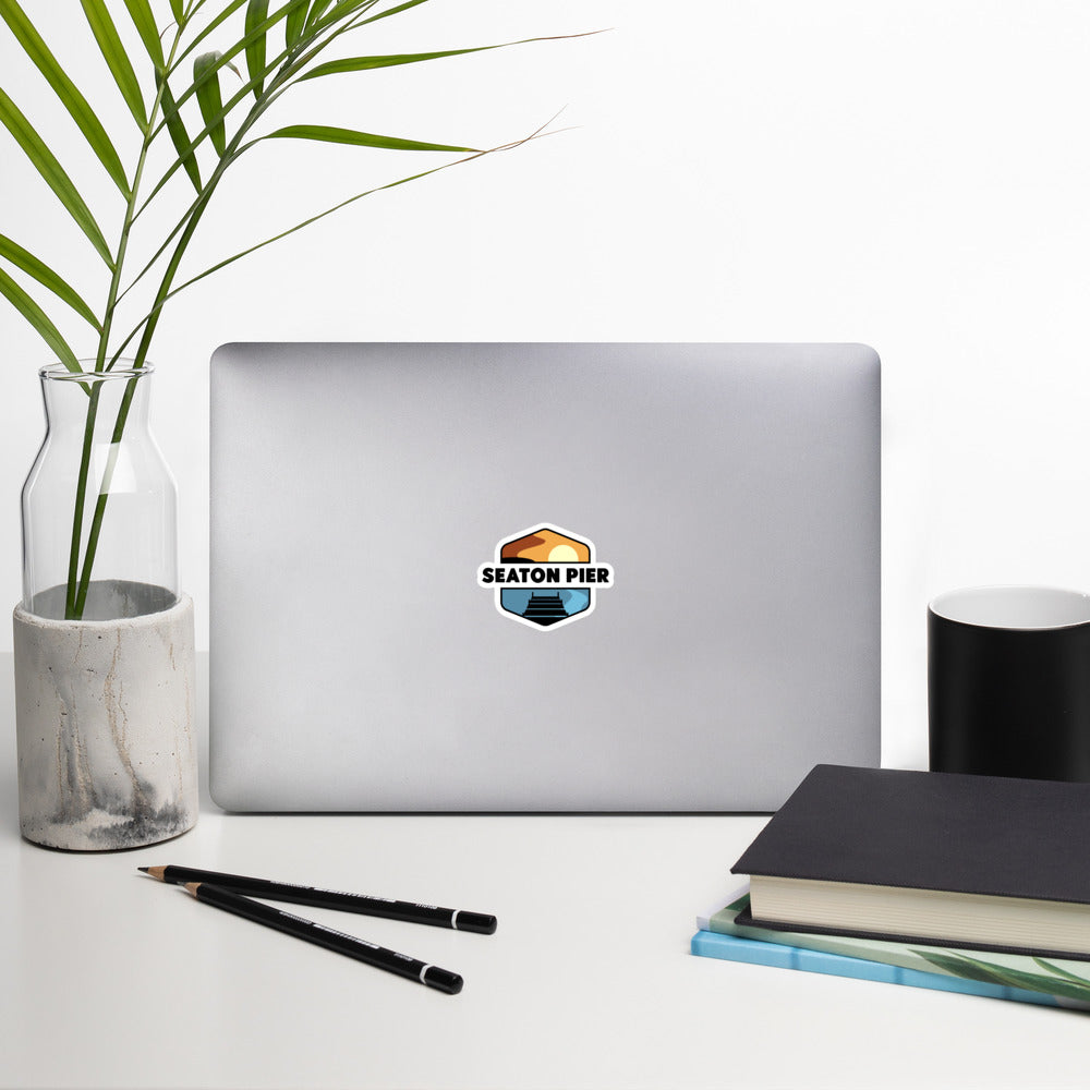 A sticker that shows a colorful setting sun over an ocean pier. With the words Seaton Pier in the middle, printed on a sticker that is 4x4 inches