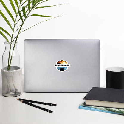 A sticker that shows a colorful setting sun over an ocean pier. With the words Seaton Pier in the middle, printed on a sticker that is 4x4 inches