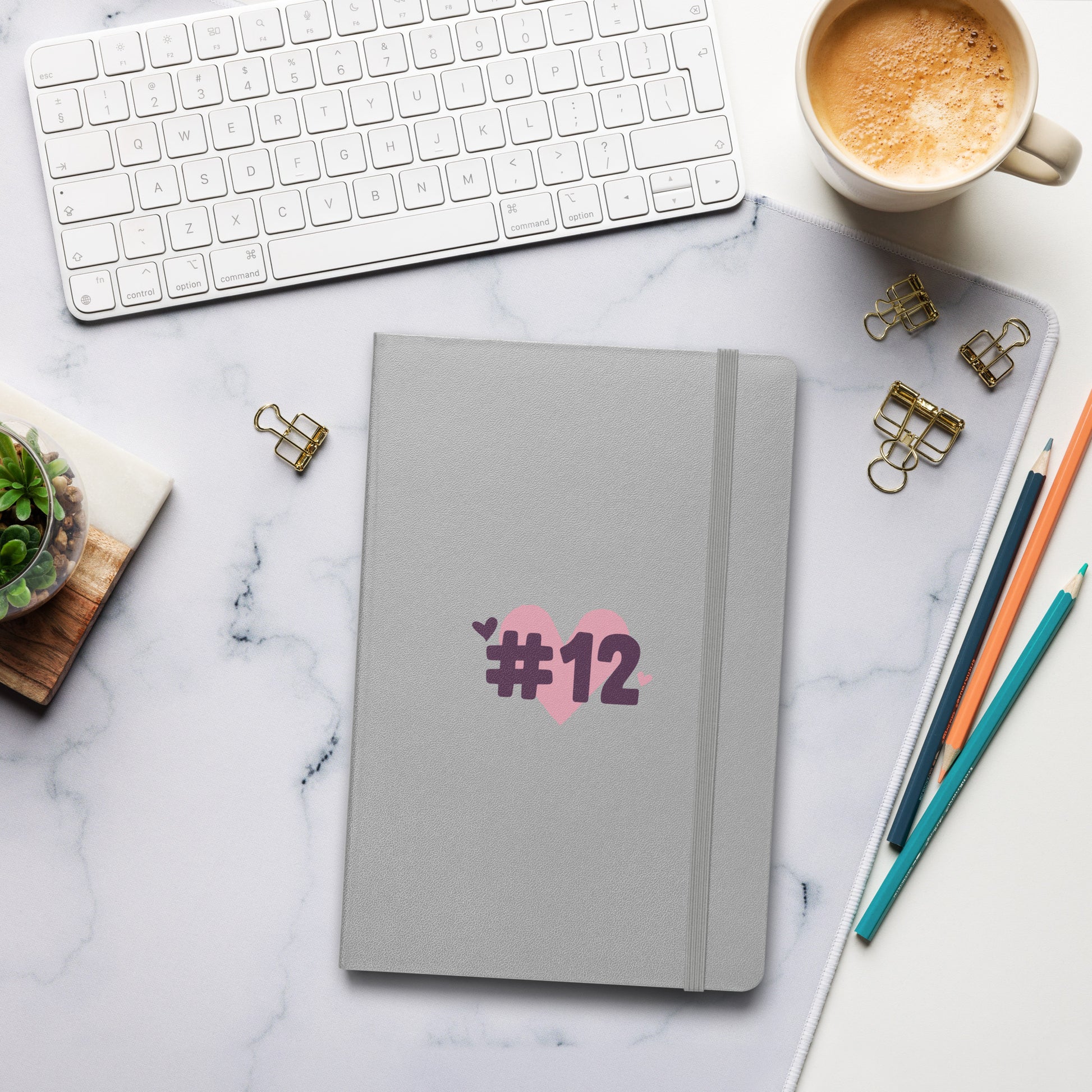 a silver notebook with the I Heart #12 Logo. The Number 12 Logo is a large pink heart with #12 in black written over the heart.