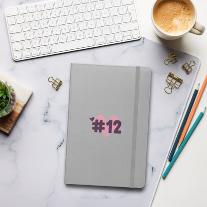 a silver notebook with the I Heart #12 Logo. The Number 12 Logo is a large pink heart with #12 in black written over the heart.