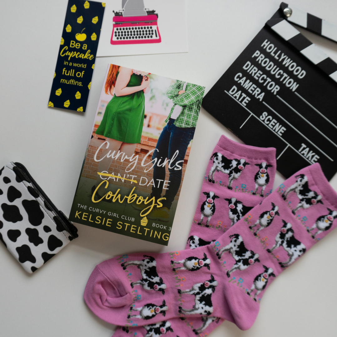 stickers, socks and a bookmark  sit next to a paperback.  The book's title is Curvy Girls Can't Date Cowboys, The Curvy Girl Club Book 3, by author Kelsie Stelting